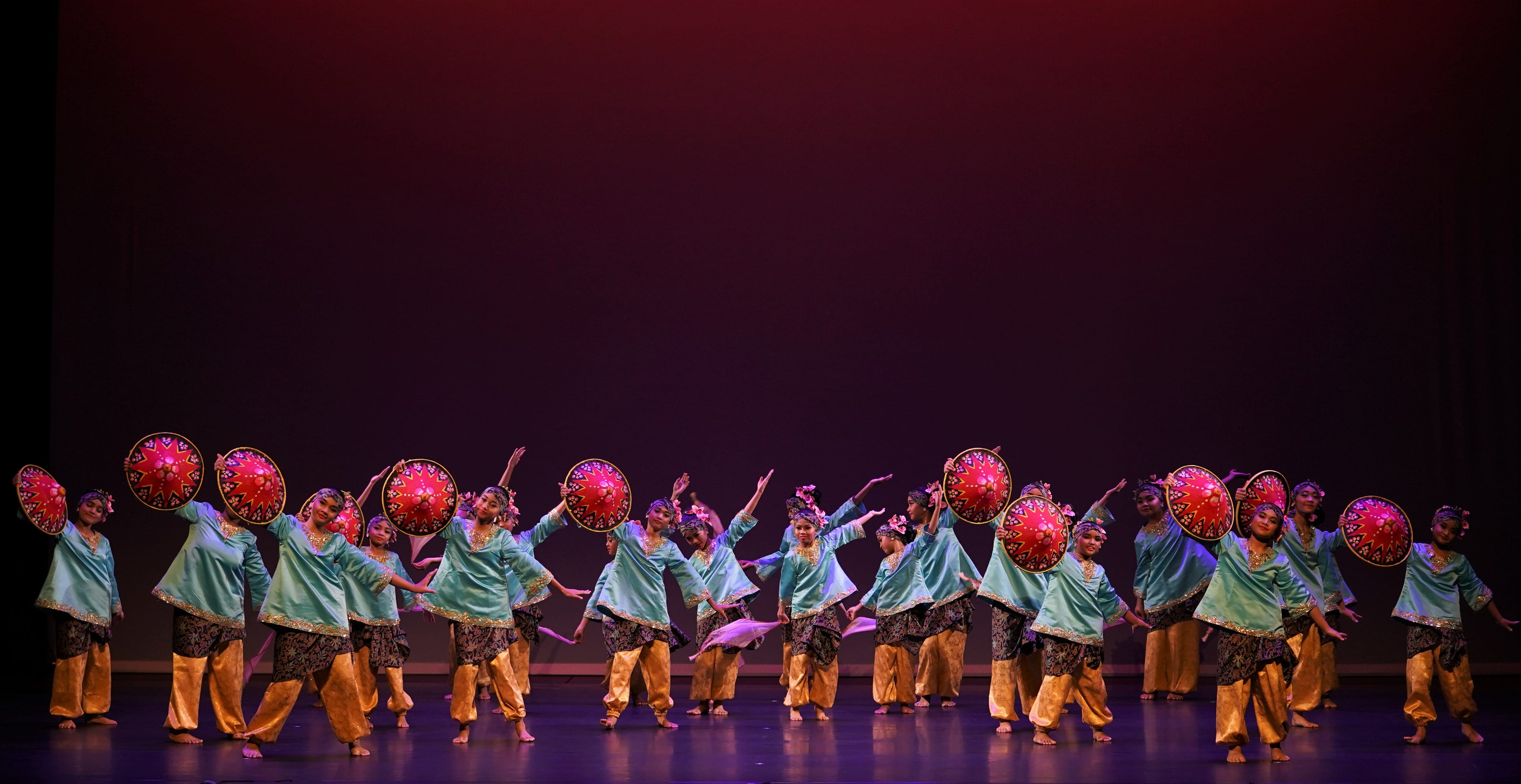 A large group of dancers in teal tops and gold pants perform on stage holding decorated red circular props.
